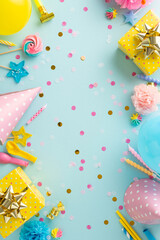 Joyful festivity creativity. Vertical top view of table set with sweets, wrapped gifts, party hats, noise-maker, candles, straws, balloons, confetti on soft blue backdrop with available space for text