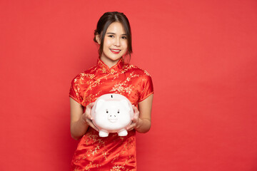 Fototapeta na wymiar Asian woman wearing red traditional cheongsam qipao dress showing and holding white piggy bank isolated on red background, Business and financial concept