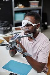 Fototapeta na wymiar Vertical portrait of Black man wearing magnifying glasses fixing quadcopter drone at workstation in repair shop