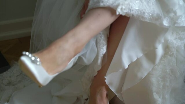 Bride put on wedding shoes. White luxury bridal footwear. Young woman in long luxury bridal gown.
