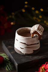 Hot winter drink in a clay mug. Close-up of hot chocolate with cinnamon stick, gingerbread house. Winter cozy still life with cocoa, Christmas decorations, fir branch and lights. © Lizaveta