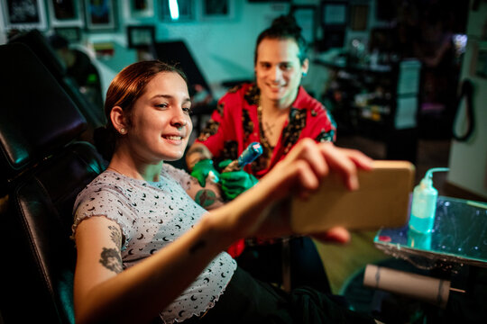 Young woman taking selfie while getting tattoo in studio