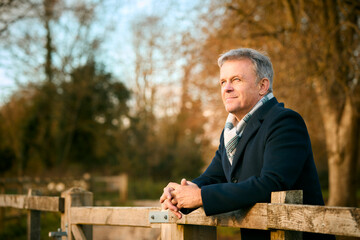 Fototapeta premium Smiling Senior Man Leaning On Wooden Fence On Walk Through Autumn Park Or Countryside