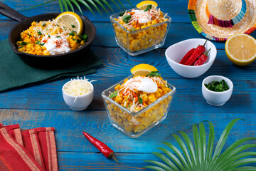 Esquites, grilled corn, served with cheese, sour cream and chili powder in glass cup on blue table. Mexican street food meal. Elote corn cup