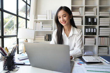 Obraz na płótnie Canvas Cheerful business lady working on laptop in office, Asian happy beautiful businesswoman in formal suit work in workplace. Attractive female employee office worker smile.