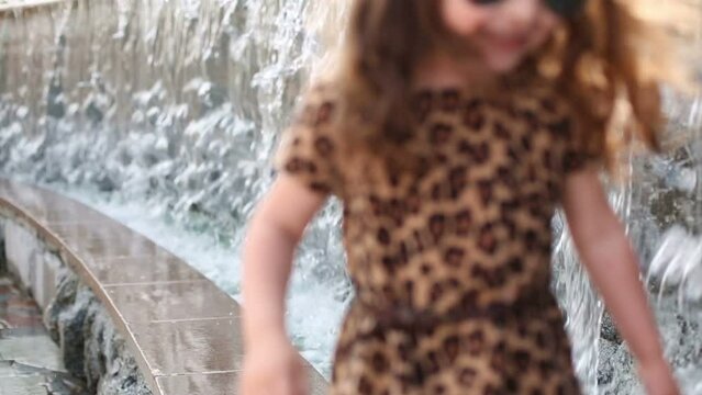 Little barefoot girl in sunglasses and dress walks near fountain