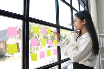 Confident focused businesswoman writing ideas or tasks on sticky papers on glass wall, female team...