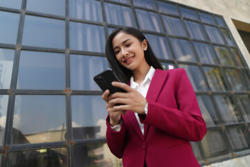 Smiling young Asian business woman wearing suit standing on urban escalator using applications on cell phone, reading news on smartphone, fast connection, checking mobile apps outdoors.