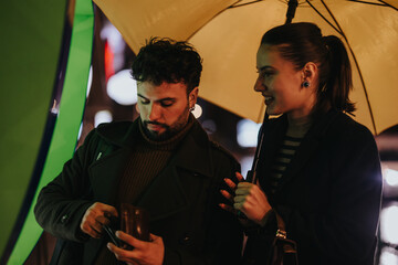 A couple of stylish people using an ATM machine on a rainy day, holding an umbrella while having a...