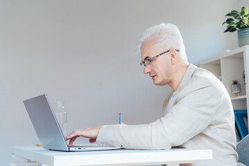 Concentrated grey hair middle-aged man making notes while working on laptop at work place in home...