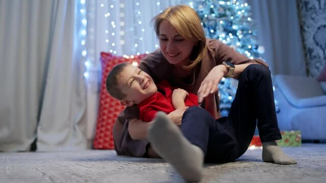 A mother tickles her child against the background of a Christmas tree. The boy laughs and squirms. Happy fun moments together. High quality 4k footage