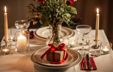 holidays, romantic date and celebration concept - close up of festive table serving for two with gift box, flowers in vase and candles burning at home on valentine's day