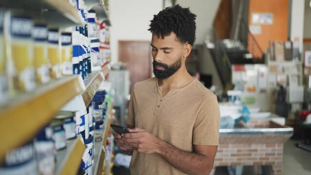 Online shopping list. Serious Bearded black man with cell phone choosing products in the shelf at hardware store. 