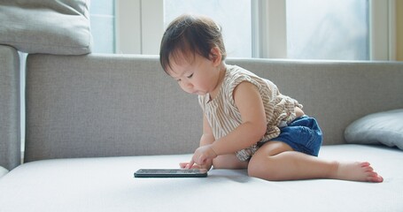 Happy little Asian child girl looking using and touch mobile phone screen technology device sitting on couch at home. toddler smiling funny time to use mobile phone, She is funny and emotional