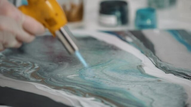 Female artist using gas burner warming colored art epoxy resin on canvas mixing colors in art studio workshop, creative woman, trendy modern hobby creating beautiful sea waves acrylic paintings