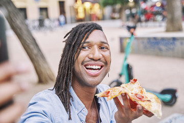 attractive man with braids holding a slice of pizza in his hand while taking a photo with his smart...