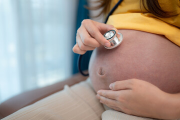 Asian pregnant woman listening to baby heartbeat with stethoscope at home