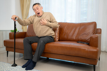 With heart disease symptoms, a senior guy sits on a sofa with a walking stick in his living room at home.
