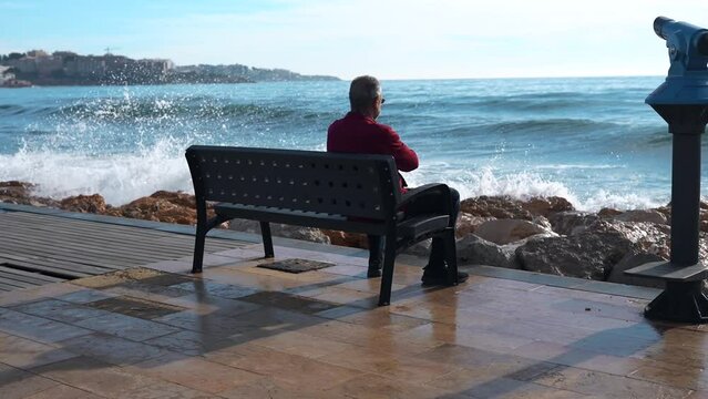 A lonely elderly man sits on a bench and looks at the sea.