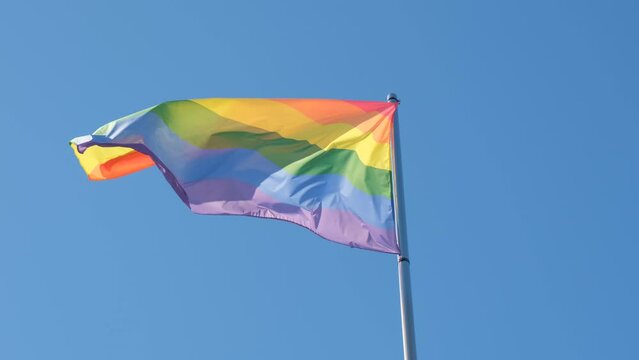 A rainbow flag, a symbol of the LGBT community, fluttering in the wind against a blue sky.