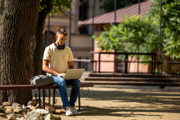 Obraz premium Short-haired young guy sitting under the tree and working on a laptop