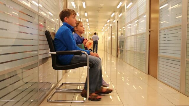 Boy and girl sit on chairs and swing legs in office corridor