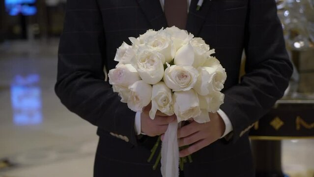 Groom in a black suit with a bouquet of flowers of white and pink roses and peonies with ribbons. A man businessman carries a gift to his love woman. Romantic day.
