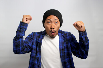 Surprised young Asian man, dressed in beanie hat and casual shirt, is celebrating success and...