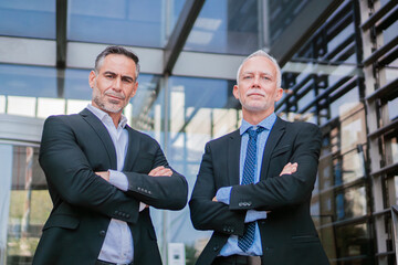 Two serious and mature businessmen looking at camera wearing classic suits standing with arms crossed gesture showing power and leadership in the world of finance . Team of male entrepreneurs posing