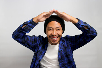 Happy Young Asian man with a beanie hat and casual clothes is making a hand gesture above his head, symbolizing a roof or home while standing against white background