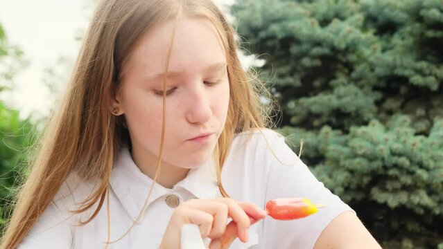 Joyful young lady enjoys frozen fruit dessert among greenery. Cheerful teenager lick lips while eating ice cream closeup