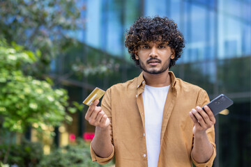 Close-up portrait of a young Indian man standing outside on the street, holding a mobile phone and...