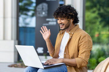 Smiling young Indian man sitting outside on a bench in the street wearing headphones and talking on a video call through a laptop, waving to the camera