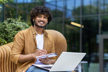 Portrait of a young Indian male student sitting outside a university campus on a bench wearing headphones, holding a notebook and studying online on a laptop. Looking and smiling at the camera