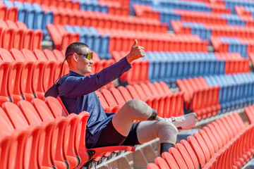 Sport man athlete prosthesis legs sit on chair of amphitheater look forward with show thumbs up in...