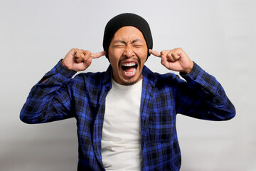 Angry young Asian man, in a beanie hat and casual outfit, is closing his eyes and covering his ears...