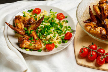Farfalle pasta with grilled chicken wings on a white plate decorated with cherry tomatoes and green onions next to a dish with wings. Medium plan.