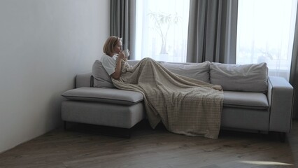 A young woman drinks hot tea while lying on the sofa. A woman on the sofa, covered with a blanket,...