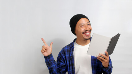 An excited young Asian student, dressed in a beanie hat and casual clothes, is smiling at the camera while pointing aside at an empty copy space, standing against a white background