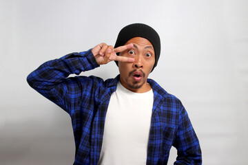 Excited young Asian man, wearing a beanie hat and casual shirt, is making a peace gesture towards camera with a V sign while standing against a white background