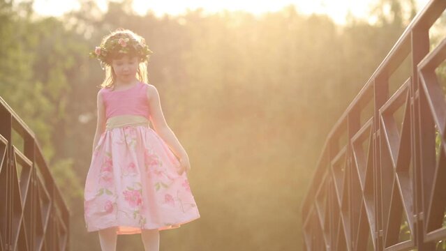 Little pretty girl in dress and wreath dances on bridge in park