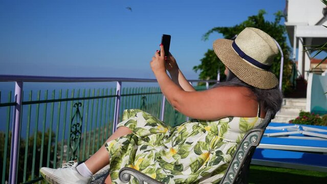 Side view of female tourist in hat and sunglasses using smartphone in Amalfi, taking pictures, shooting video sitting chair on background of beautiful seascape, on summer sunny day, slow motion.