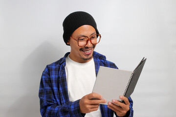 An excited young Asian student, wearing glasses, a beanie hat, and casual clothes, is immersed in reading a book while standing against a white background