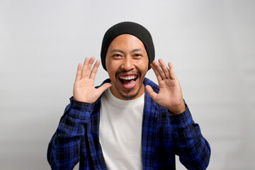 Asian man, dressed in a beanie hat and casual shirt, brings his hand near his mouth as if sharing a...