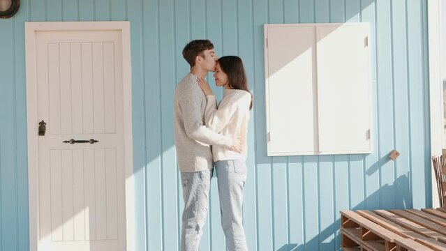 A tender moment of a young couple standing by a light blue beach house on a sunny day, sharing a kiss and expressing their love symbolizing moving into new house