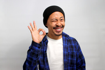 Excited young Asian man, dressed in a beanie hat and casual shirt, blinks at the camera while displaying the OK sign gesture. He appears satisfied, indicating approval and giving a positive review.