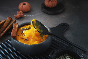 Delicious pumpkin soup with heavy cream, sesame seeds and pumpkin seeds on a dark rustic wooden table with toast. A spoon scoops up pumpkin cream soup