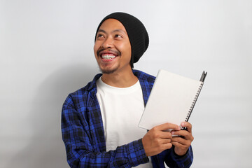 A young Asian student, dressed in a beanie hat and casual shirt, gazes at an empty space, pondering education and the future while holding an open book, standing against white background.