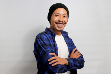 Confident young Asian man, clad in a beanie hat and casual outfit, stands with folded arms, wearing a confident smile as he gazes at the camera while standing against a white background