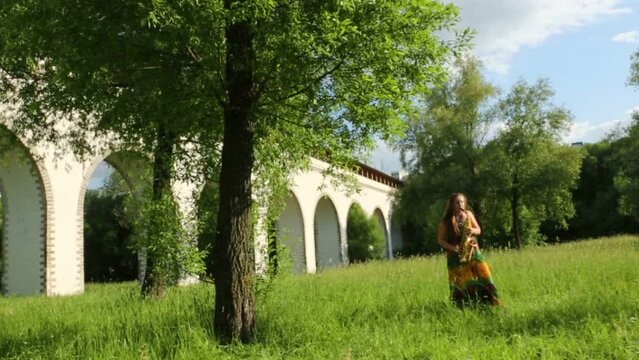 Girl stands among grass and plays saxophone near aqueduct